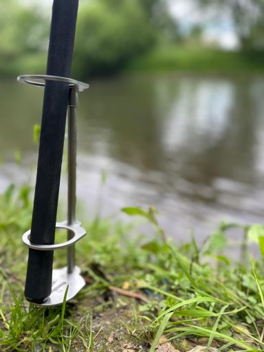 Ein Stab mit einem Metallring steht am Ufer eines ruhigen Gewässers, umgeben von Gras.