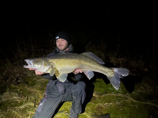 Mann hält einen großen Zander bei Nacht, trägt eine dunkle Jacke und eine Mütze.