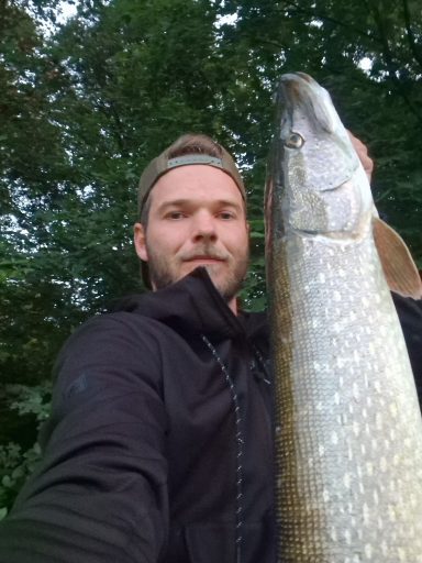 Junger Mann hält einen großen Hecht in der Hand, Hintergrund mit Bäumen.
