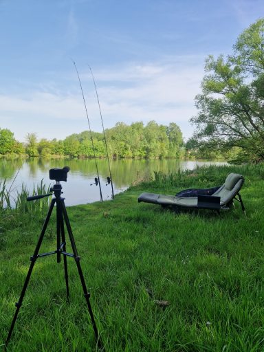 Anglerplatz am Wasser mit Kamera, Ruten und Liegestuhl in grüner Umgebung.