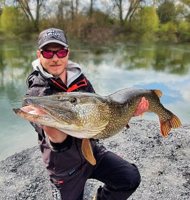 Angler zeigt einen großen Hecht am Wasser, umgeben von grüner Natur.