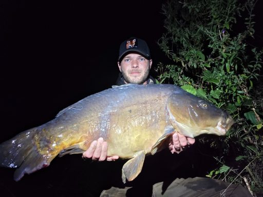 Mann hält einen großen Karpfen in der Nacht, umgeben von Bäumen.